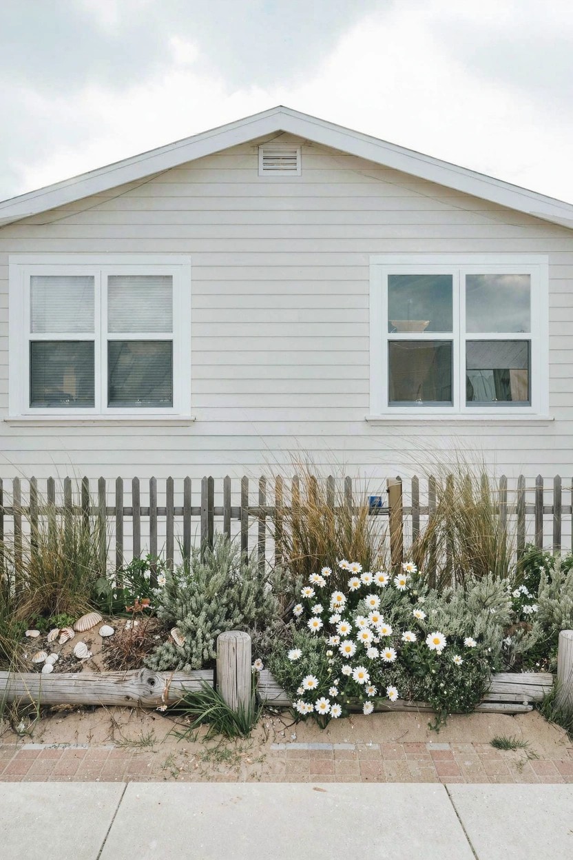 A light gray house with white clapboard siding, gabled roof, and two front double-hung windows, fronted by a white picket fence and a raised flower bed edged with stacked driftwood logs containing grasses, white daisies, succulents, rocks, and sand.