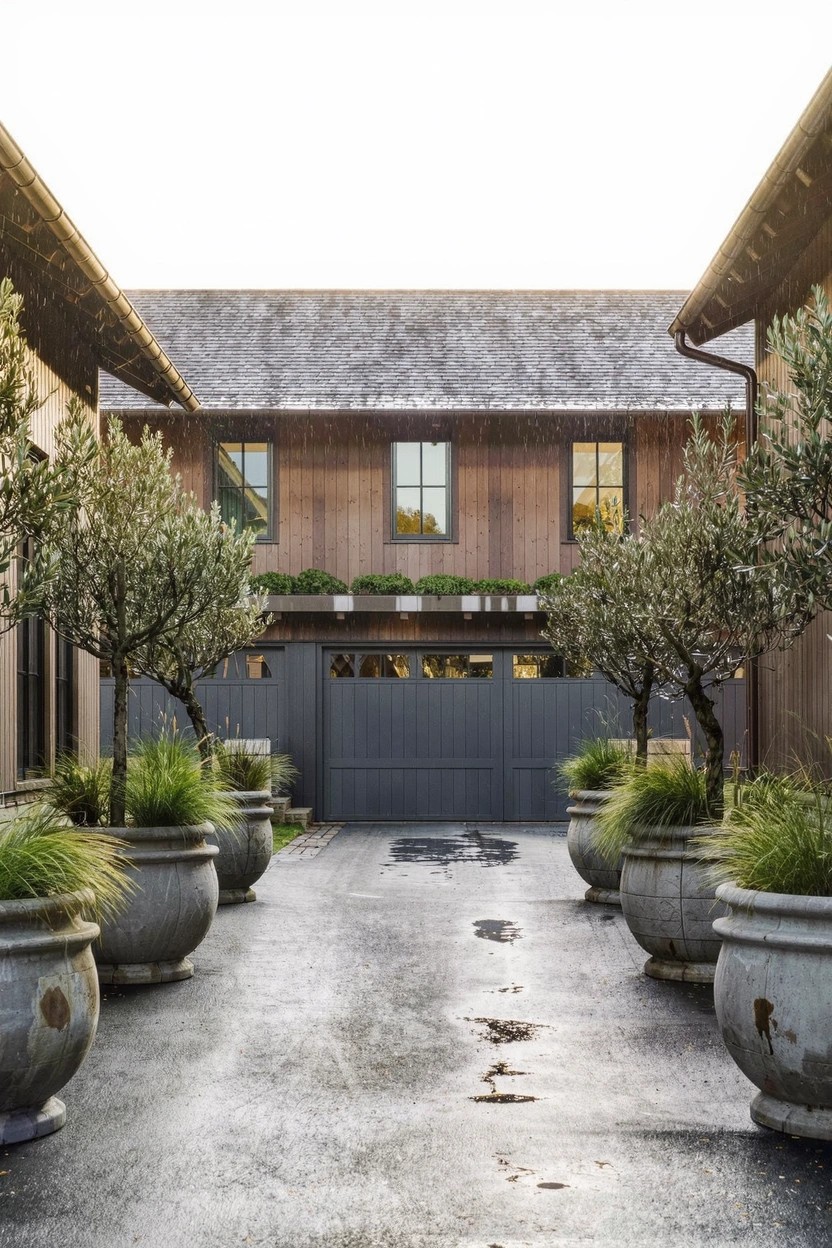 Paved pathway lined on both sides with large galvanized pots filled with grasses and flanked by olive trees, leading to gray garage doors on a cedar-sided house under an overcast sky.