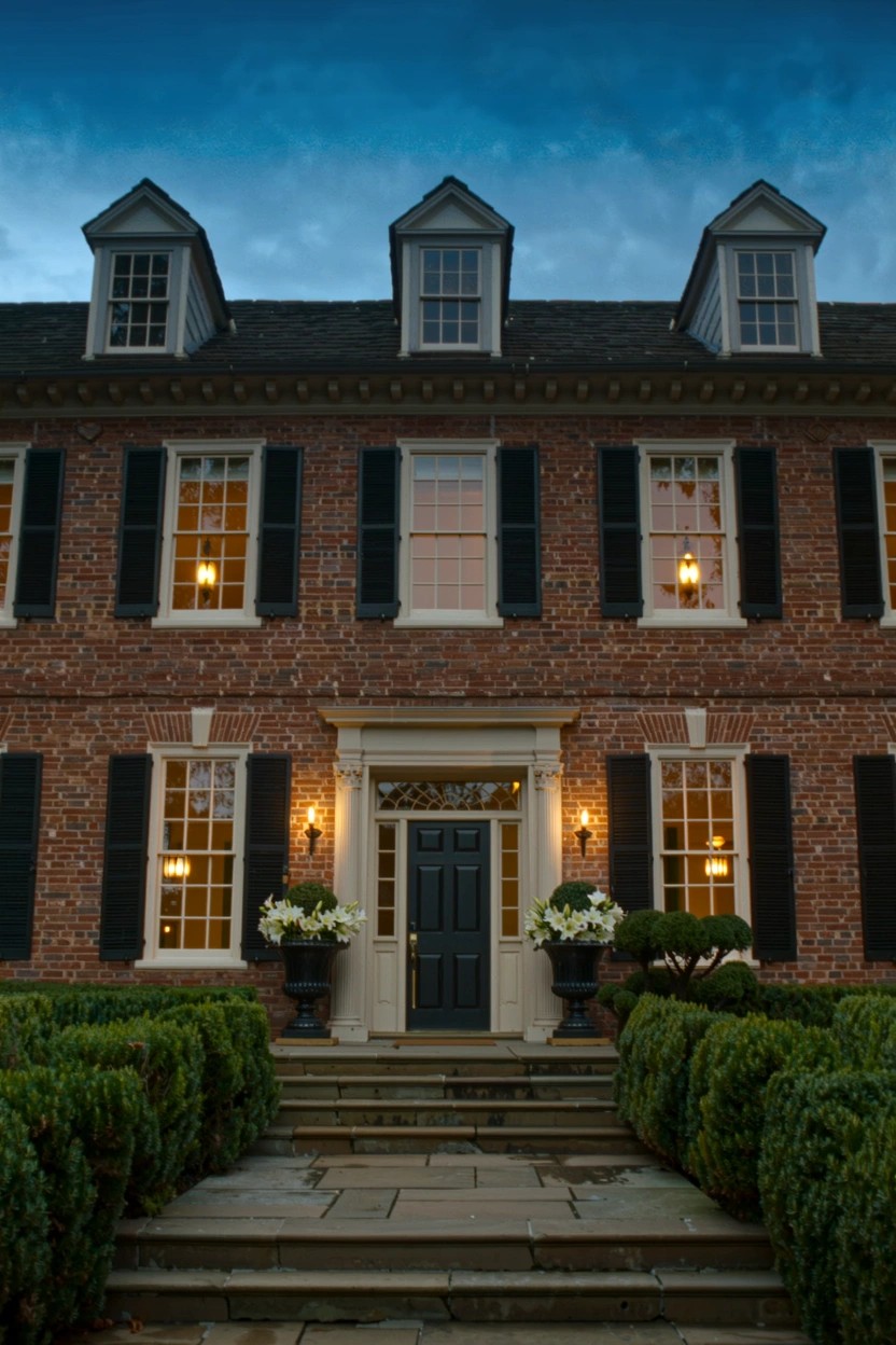 Brick colonial-style house exterior at dusk featuring large urns filled with white flowers flanking stone entry steps lined with boxwood hedges, symmetrical windows with black shutters, and a dark front door.