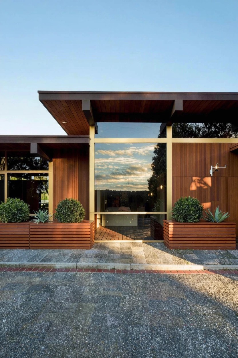 Wood-clad modern house exterior with rectangular wooden planters containing agave plants flanking the entry on a brick pathway at dusk.