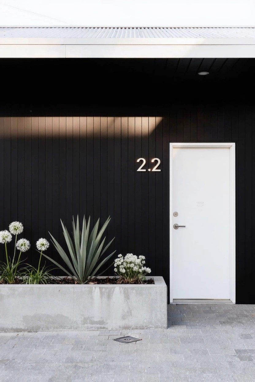 Black wood-clad exterior wall with a white door marked 2.2, next to a long concrete planter box holding agave plants and white flower clusters, on a concrete paver walkway.