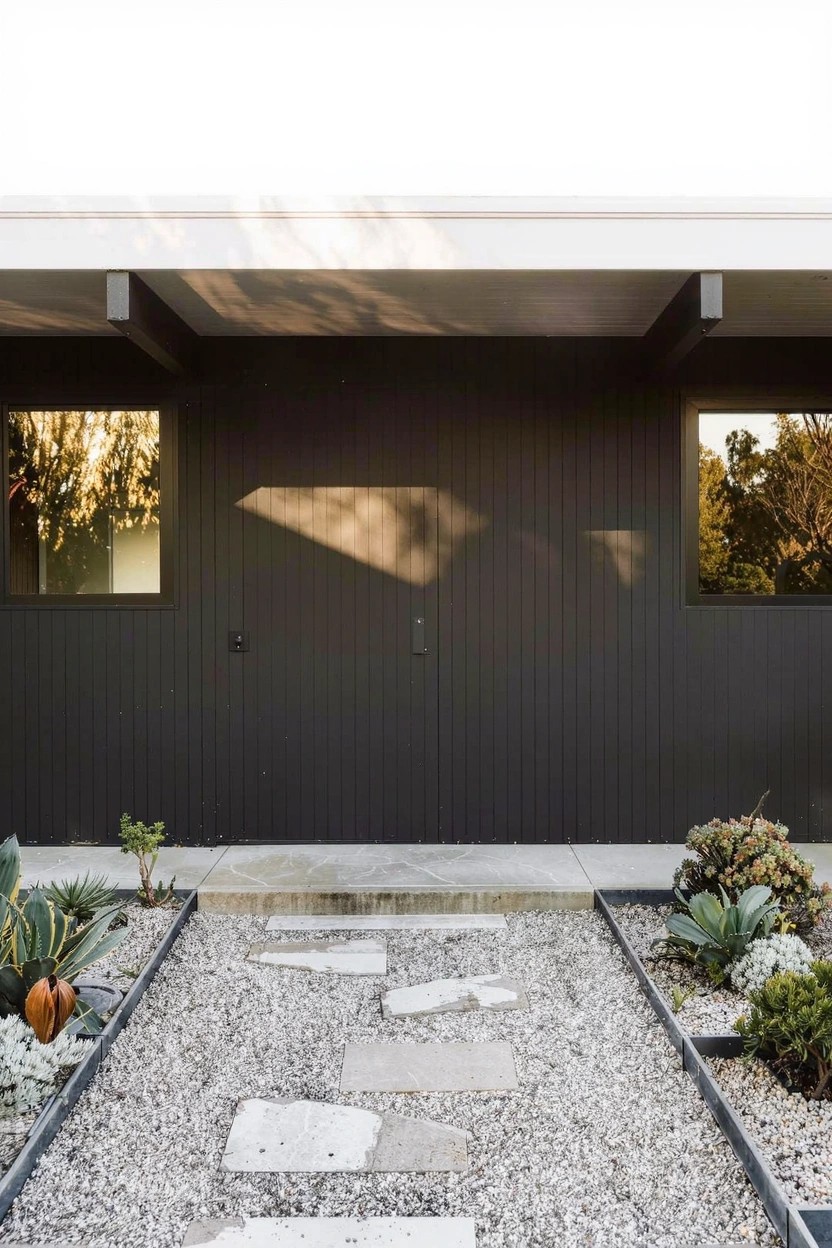 Modern house exterior featuring a dark paneled facade with two large windows, a central entry area, and a gravel pathway with irregular stone pavers leading to the door, edged by low succulents, agave, and small plants.