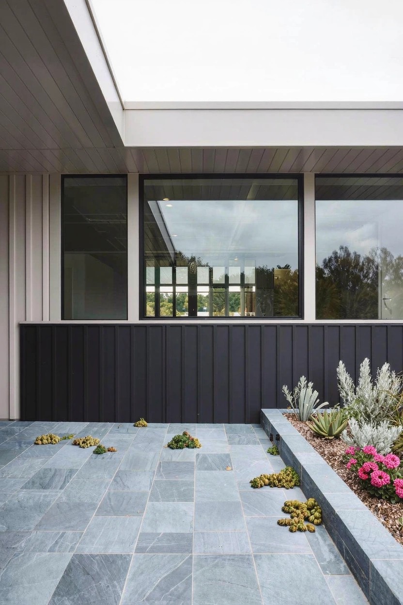 Gray slate patio corner with raised succulent bed containing agave and pink flowers, beside dark vertical wood cladding and large house windows.