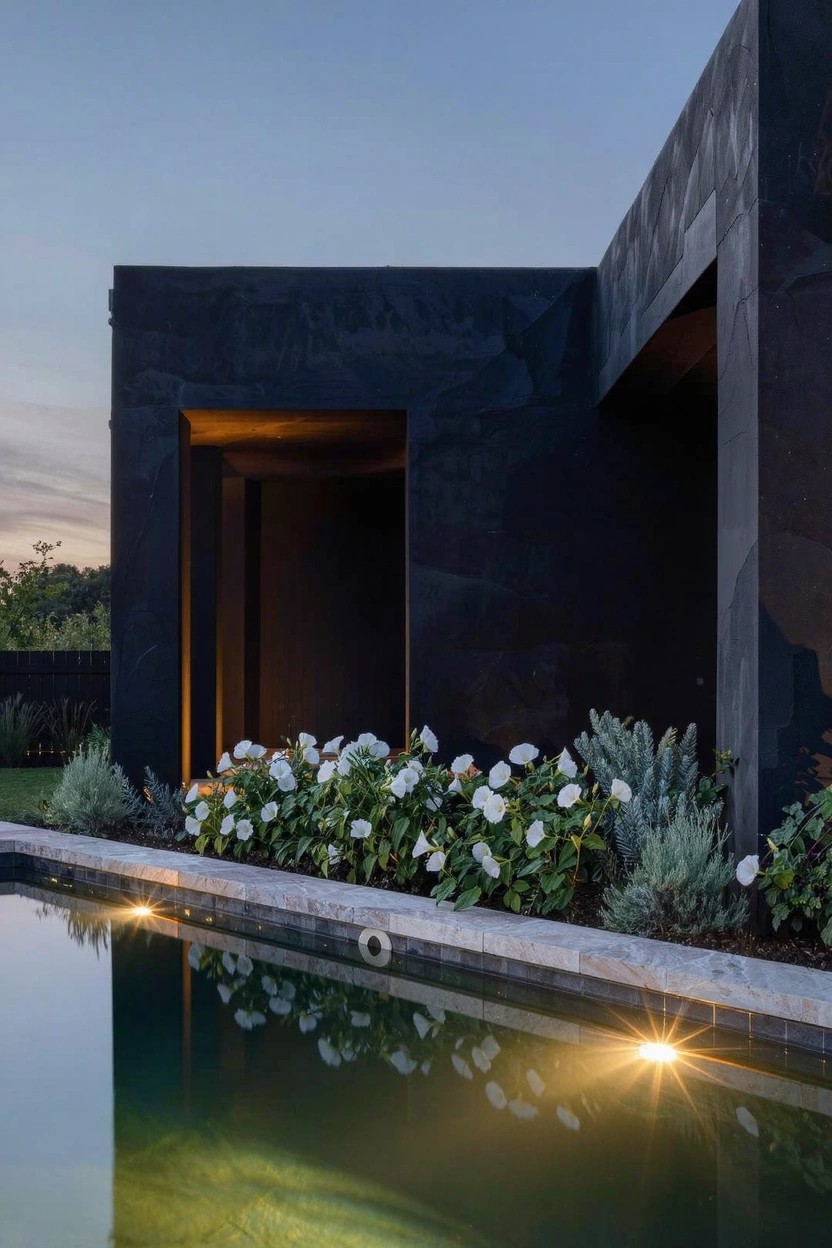 Dark angular modern house exterior at dusk next to a rectangular lit swimming pool edged by beds of white flowering shrubs and low greenery.