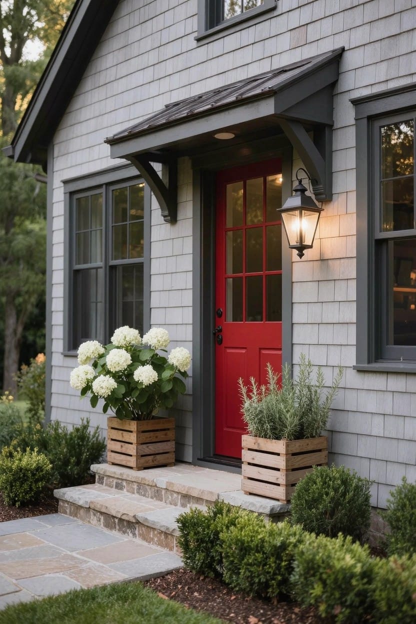 Gray shingled house exterior featuring a red front door under a small overhang, flanked by wooden crate planters with white hydrangeas and lavender pots, black lantern light, stone steps, and low shrubs.