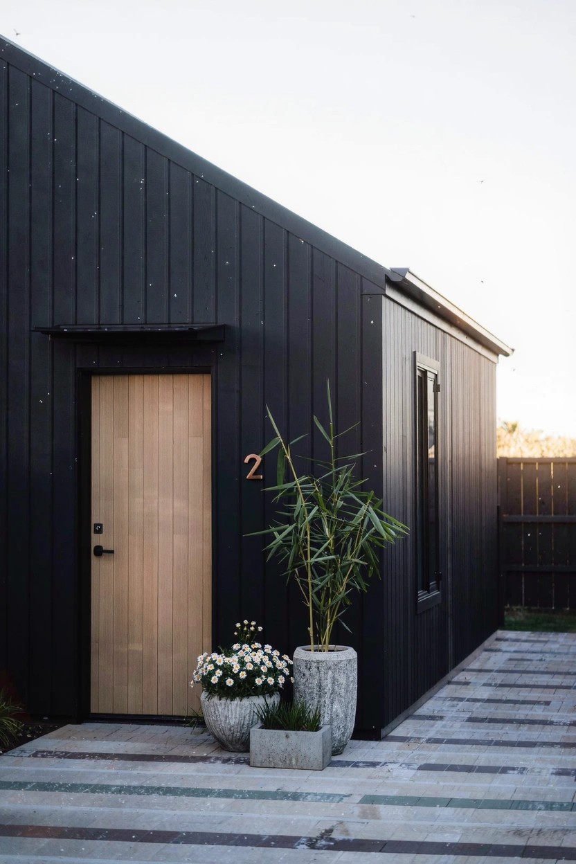 Black vertical board-and-batten shed with slanted roof and light wooden door numbered 2, flanked by two large concrete pots containing a palm plant and white flowers, on a striped concrete patio.