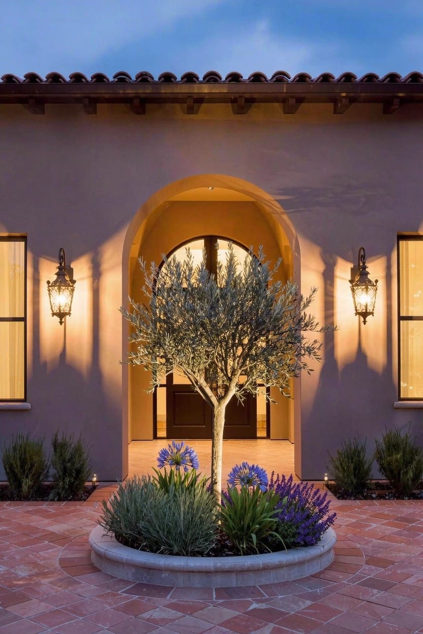 Beige stucco house with arched entry door flanked by lanterns, circular raised planter bed around a single olive tree with succulents, lavender, and agave on a brick paver walkway at dusk.