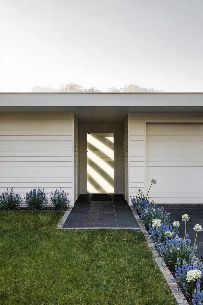 Gray board-and-batten house with white garage doors, brick-paver driveway edged by rectangular brick flower beds containing boxwood shrubs, red flowers, and other plants, plus wall lanterns.