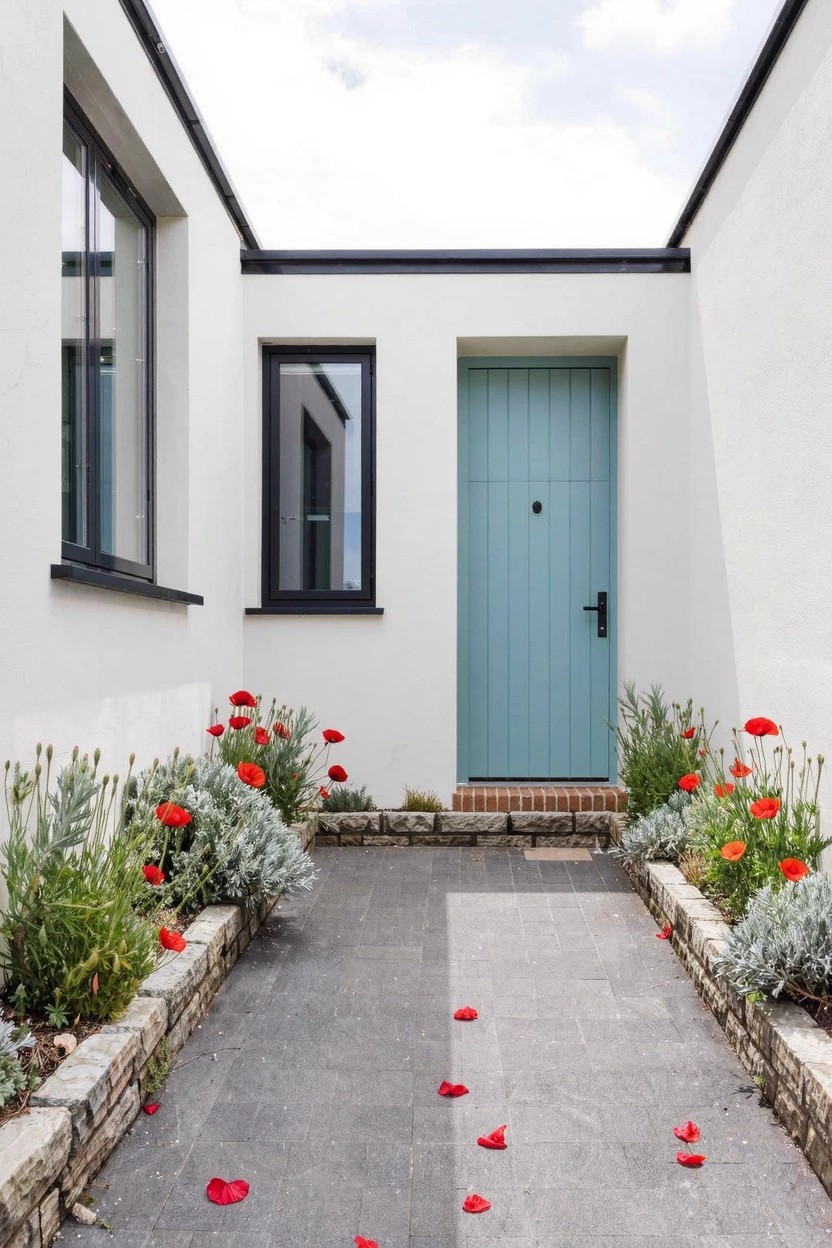 Paved gray pathway leading to a turquoise wooden door on a white-walled modern house exterior, with flower beds of red poppies, silvery foliage, and stone edging along both sides.