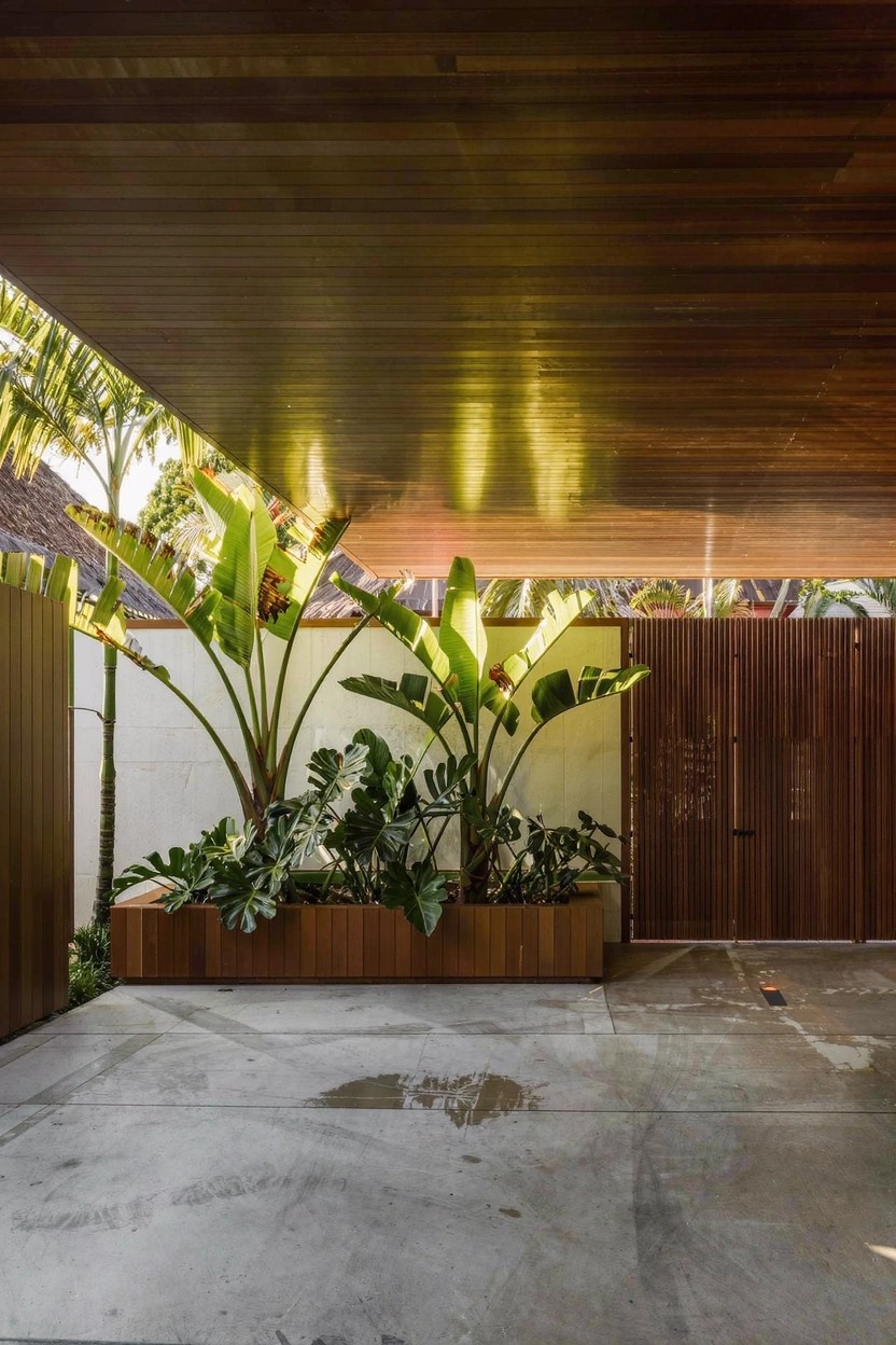 Gray modern house with stone pillars at entry, concrete driveway leading to wide stone steps bordered by flower beds containing red flowers and green plants, Japanese maple tree in front with red autumn leaves overhead.
