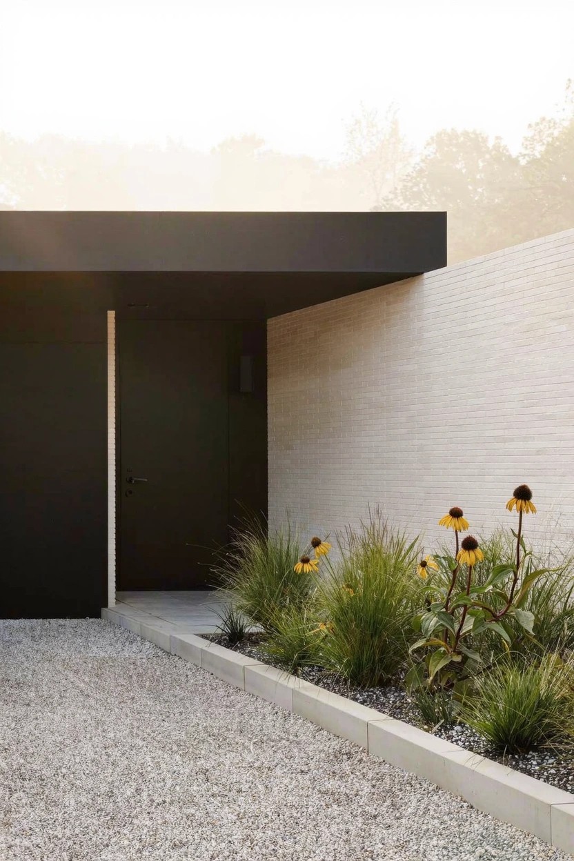 Gravel driveway bordered by a narrow flower bed with tall ornamental grasses and yellow coneflowers next to a black front door under a dark overhang on a white brick wall.