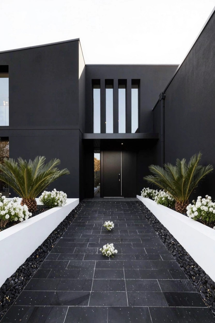 Black modern house with a straight black stone pathway to the front door, bordered by low white walls holding white flowers and potted palms.