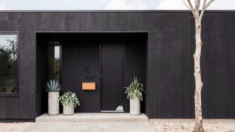 White clapboard house with gabled roof, navy blue front door flanked by two large gray pots of trailing green plants, black lanterns, black shutters, boxwood shrubs, brick steps, and gravel path.