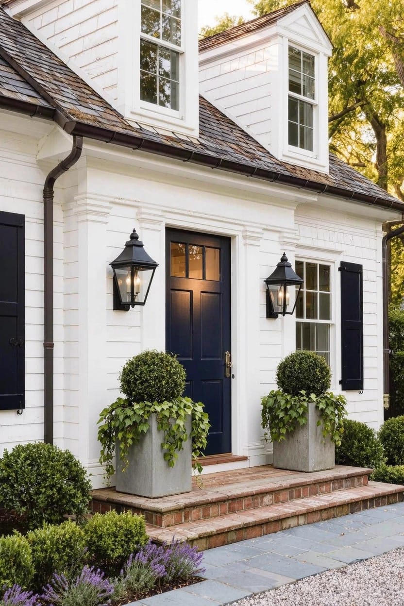 White clapboard house with gabled roof, navy blue front door flanked by two large gray pots of trailing green plants, black lanterns, black shutters, boxwood shrubs, brick steps, and gravel path.