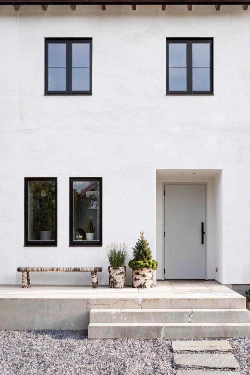White house exterior with black-framed windows, white door on concrete steps flanked by large pots of ornamental grasses, a small potted evergreen tree, and a wooden bench on gravel ground.