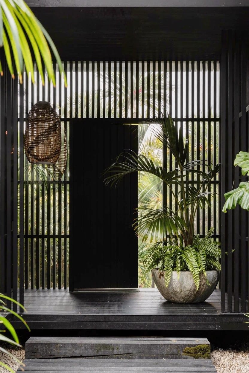 Dark timber porch deck with vertical black slats framing a closed black door, large light gray stone planter holding a large fern beside the door, hanging basket, tropical plants around, and steps leading up.
