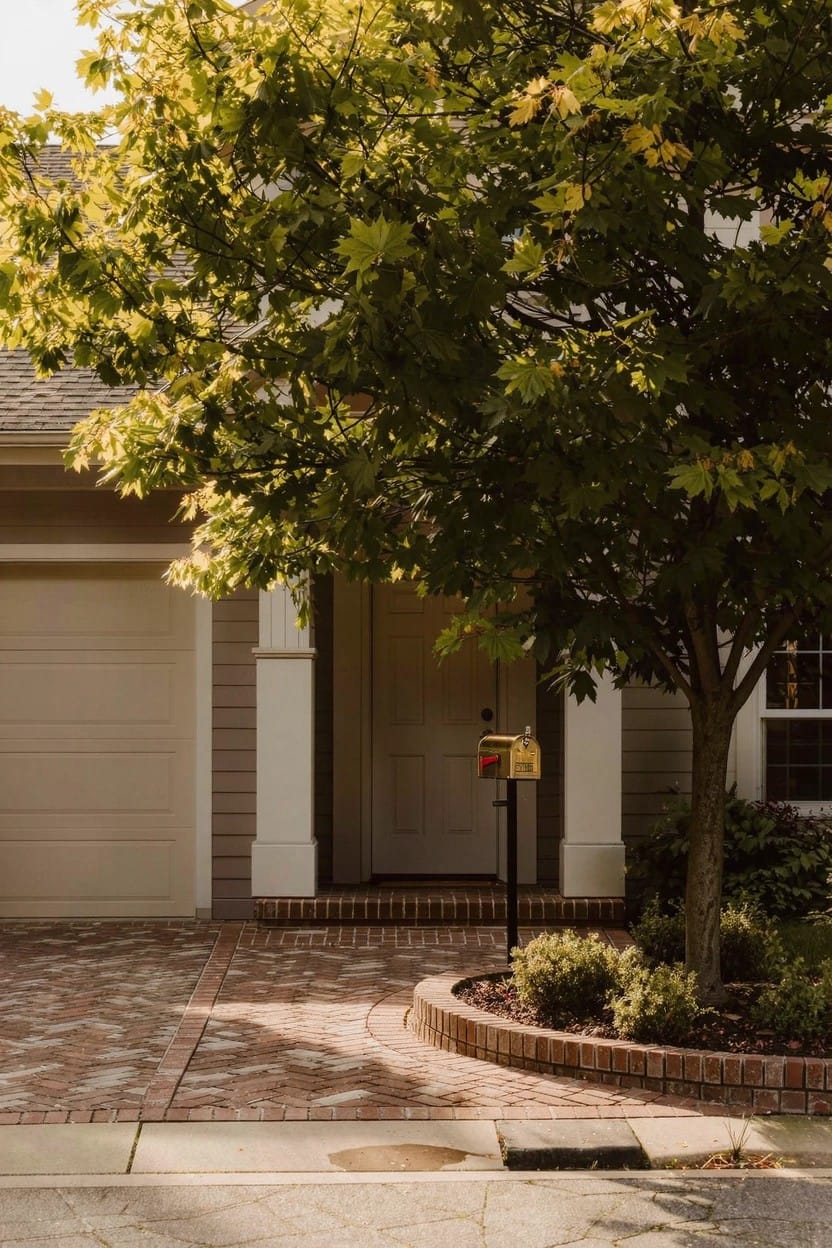 Beige house exterior with closed garage door, white-trimmed front door, large green tree, brick-paver driveway, curved brick-edged flower bed with shrubs, and mailbox on a post.