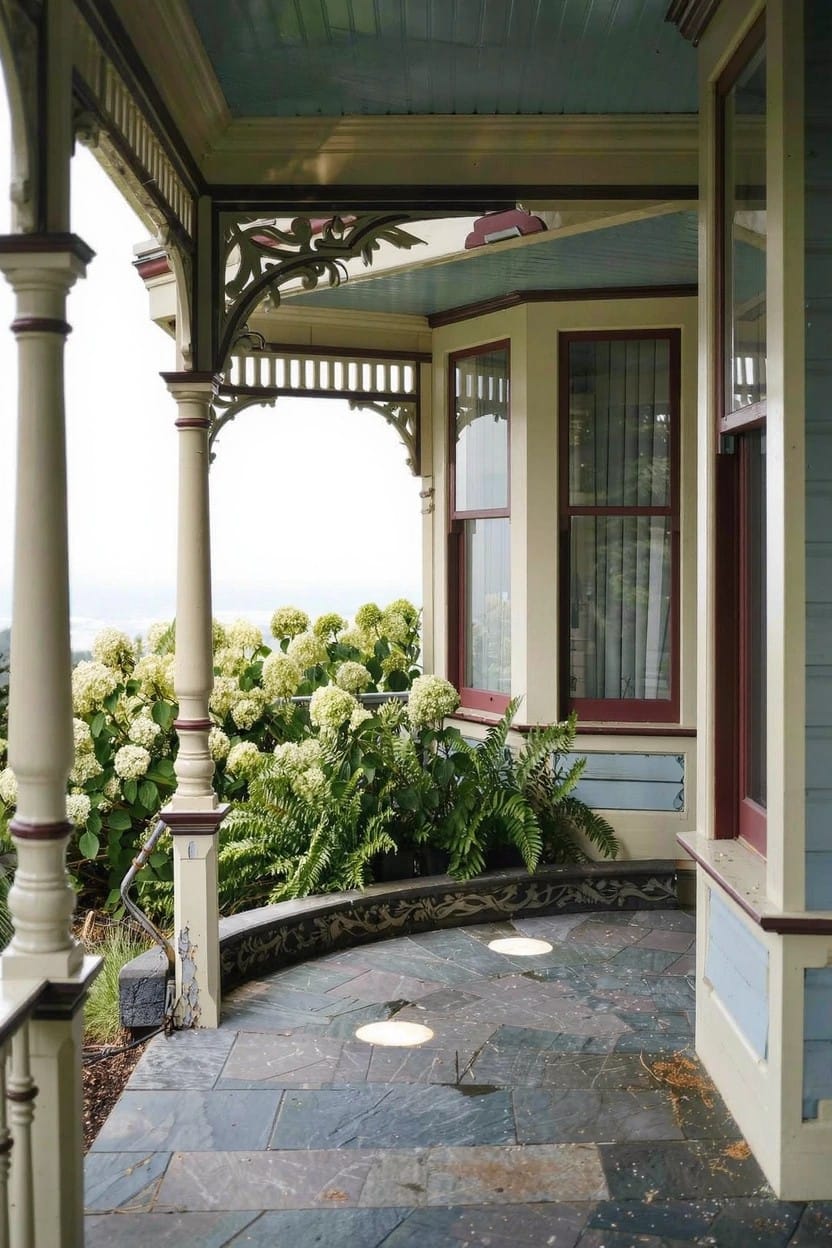 Ornate Victorian-style porch with gingerbread trim, blue ceiling, and beige siding surrounded by large white hydrangea bushes at the base, with a curved dark slate stone pathway and embedded round lights.