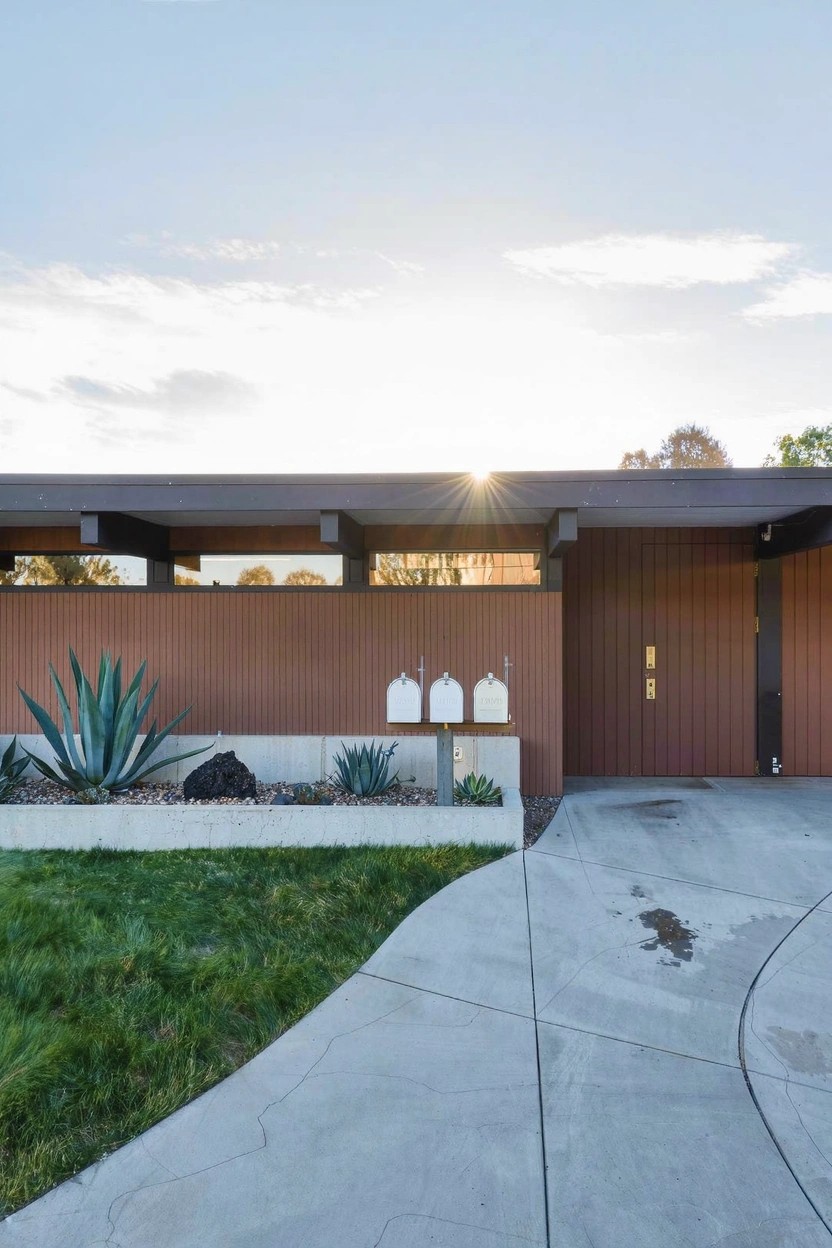 Brown horizontal mid-century modern house exterior with two white mailboxes on a raised planter bed of agave plants, rocks, and gravel next to a curved concrete driveway.