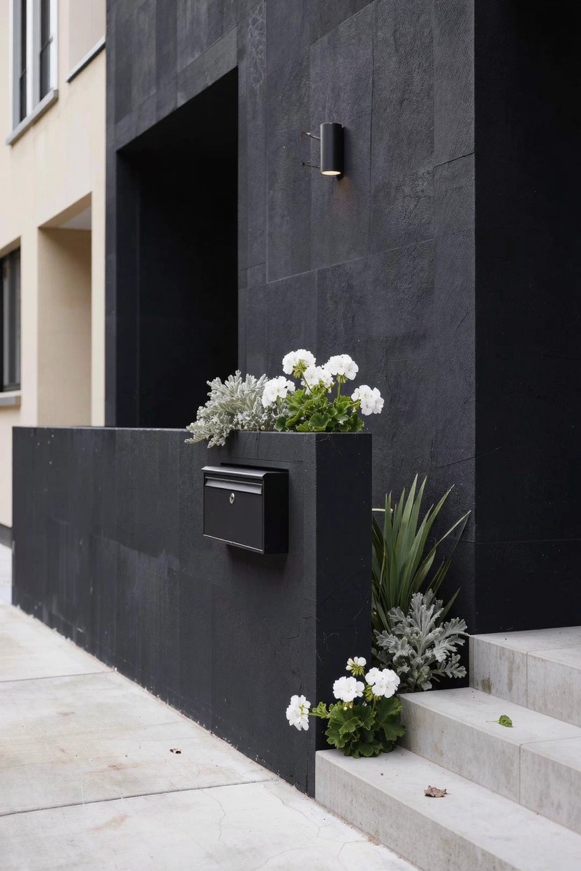 Black stone wall with integrated black mailbox and planter holding white geraniums and silver-leafed plants beside concrete entry steps on a modern home exterior.