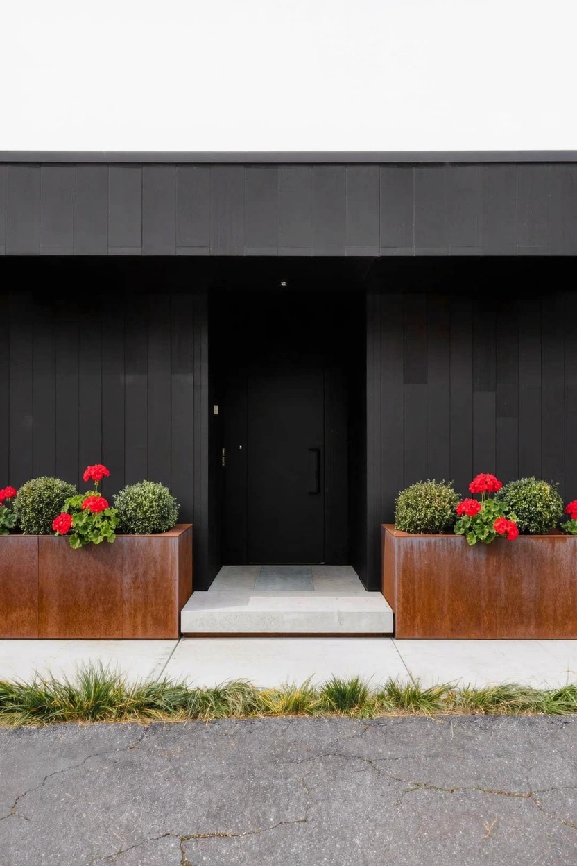 Black front door in a dark vertical-paneled facade flanked by two tall rectangular rust-colored metal planters filled with red geraniums on a concrete step.