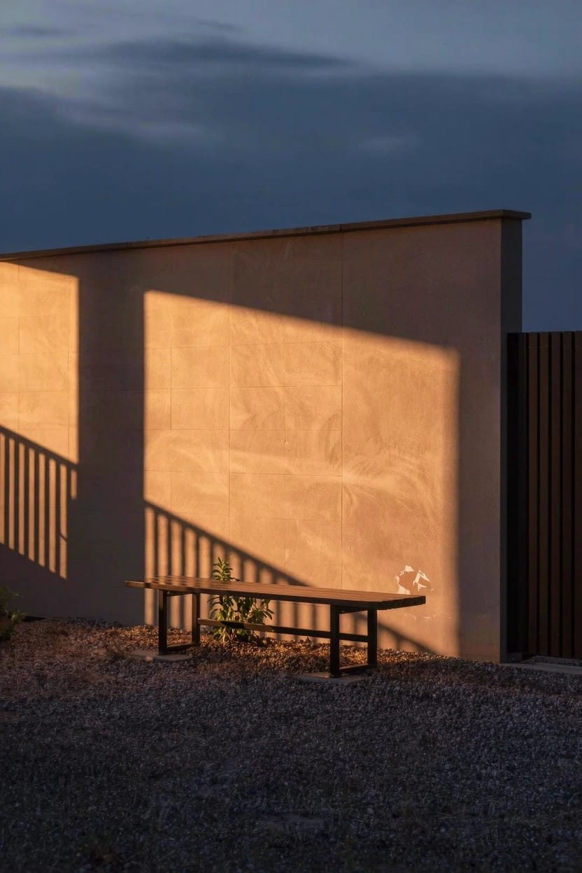 A black metal bench positioned on light gravel ground beside a beige stucco wall casting long shadows from nearby railings, with a small green plant next to the bench and a wooden fence panel in the background at dusk.