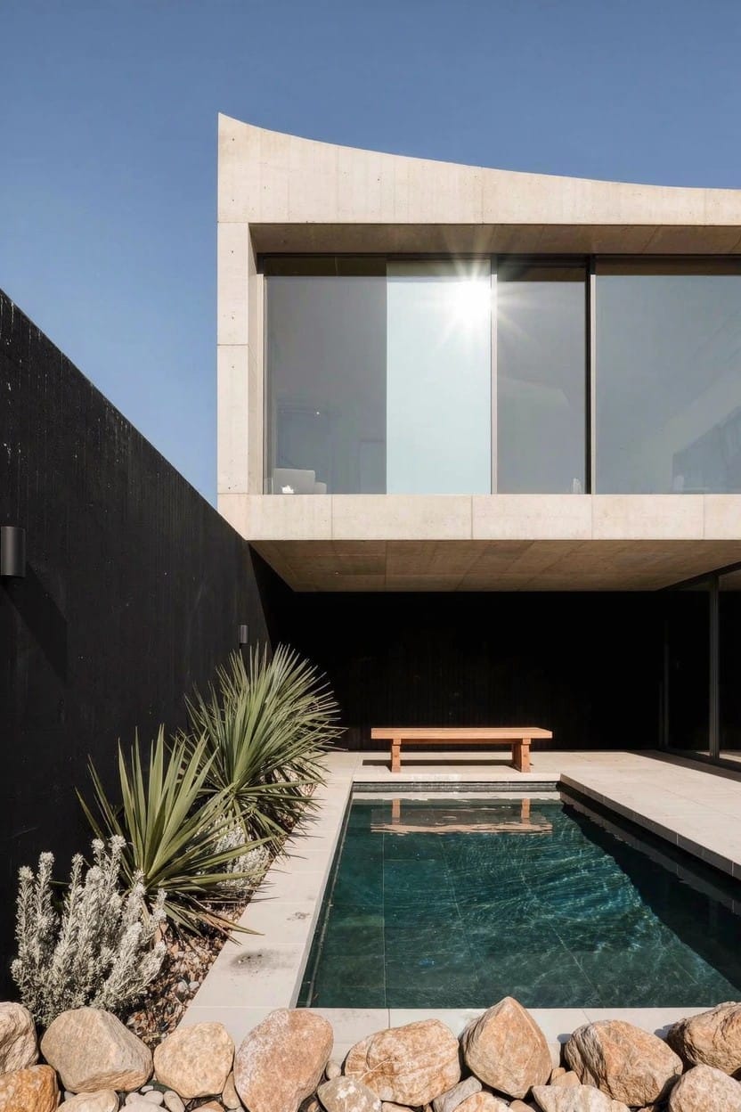 Contemporary beige concrete house with curved roofline and large glass windows beside a small rectangular pool edged with rocks and planted with agave and other succulents, a wooden bench nearby against a black wall.