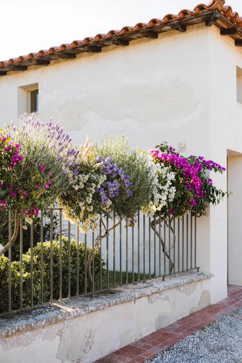 White stucco house exterior with red tile roof, black wrought-iron fence, and overflowing plants including purple lavender, white flowers, and pink bougainvillea along the fence line.