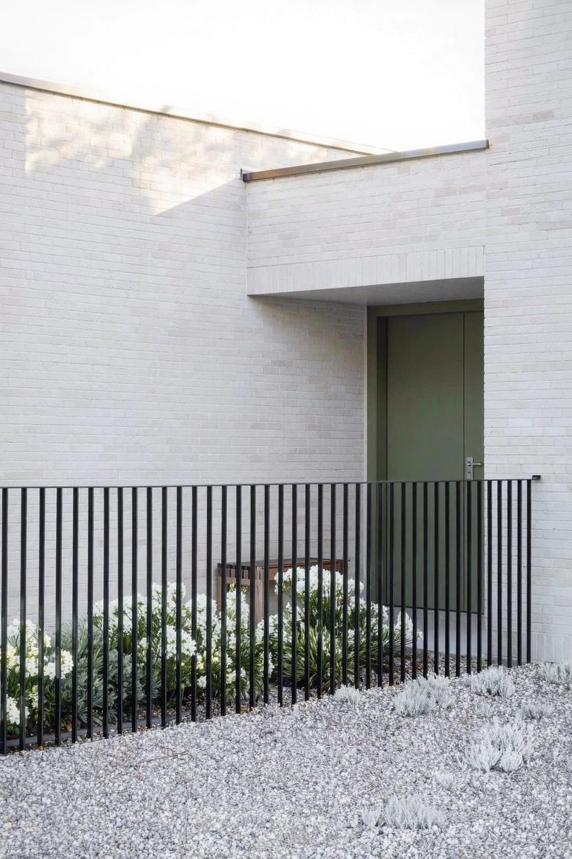 White brick house exterior with green entry door, black vertical bar fence, and low white flowering plants along the fence base on gravel ground.