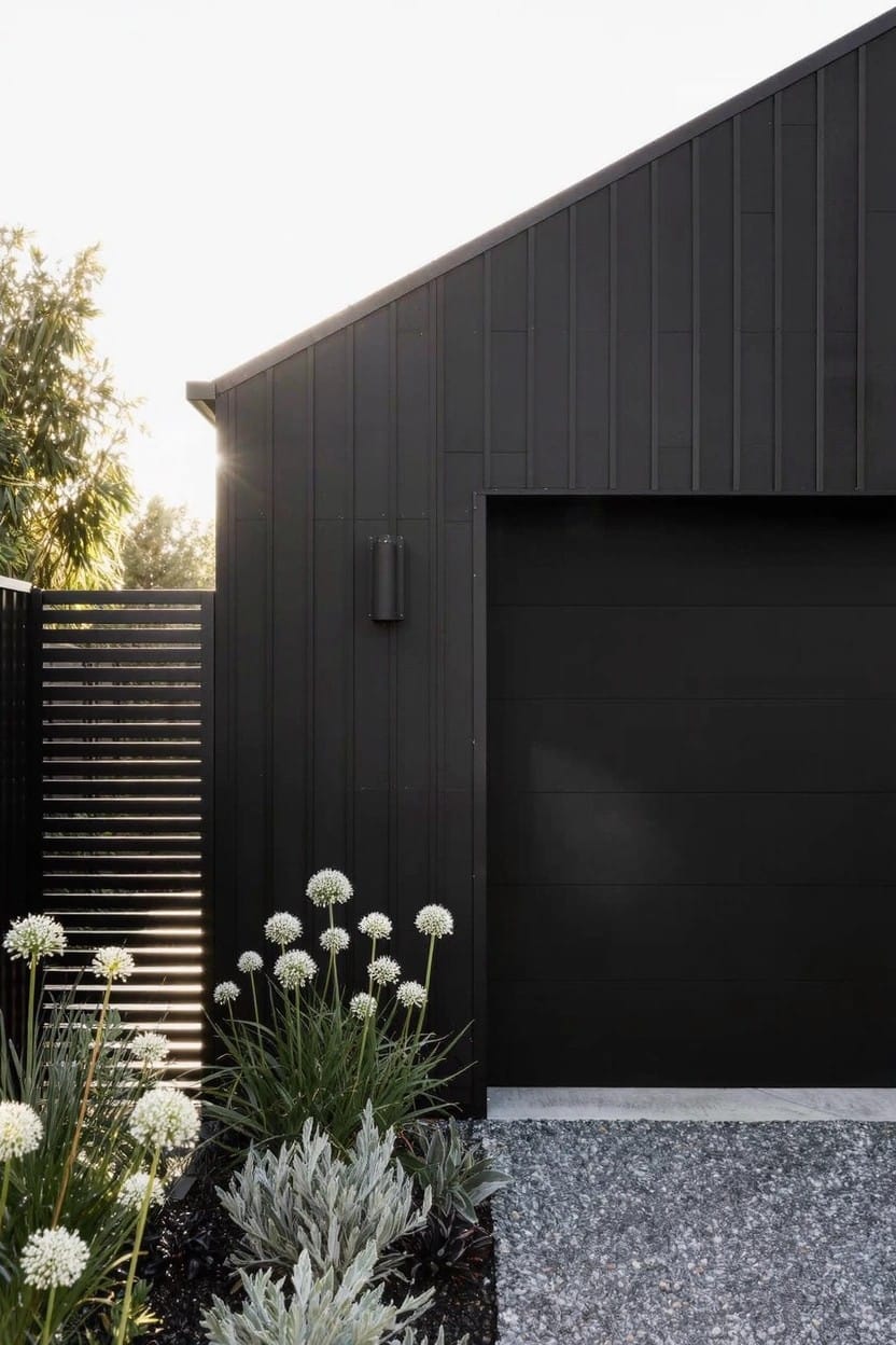 Black weatherboard garage door and wall with adjacent slatted black fence, edged by gravel and bed of white spherical flowers on tall stems mixed with strappy and silvery plants.