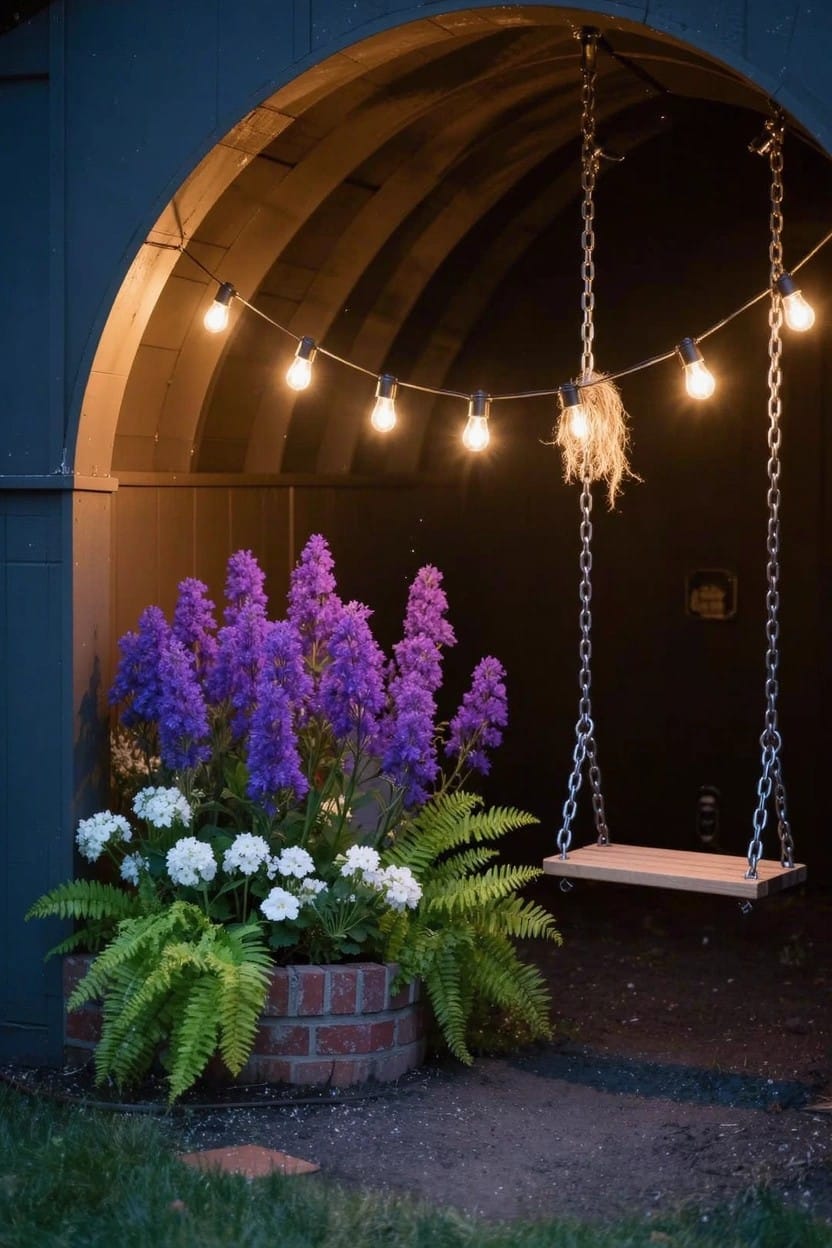 Dark arched wooden garden structure with chain-suspended wooden swing, string lights draped across the top, purple lupine flowers, white blooms, ferns in a brick planter beside it at dusk.