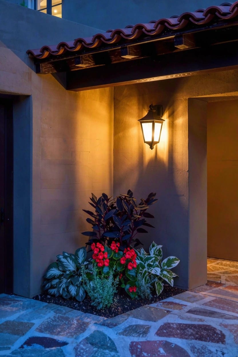 Home exterior corner at dusk showing a dark wooden garage door, glowing wall lantern, stone pathway, and shady flower bed with red flowers, dark strap-like leaves, variegated plants, and low groundcovers.