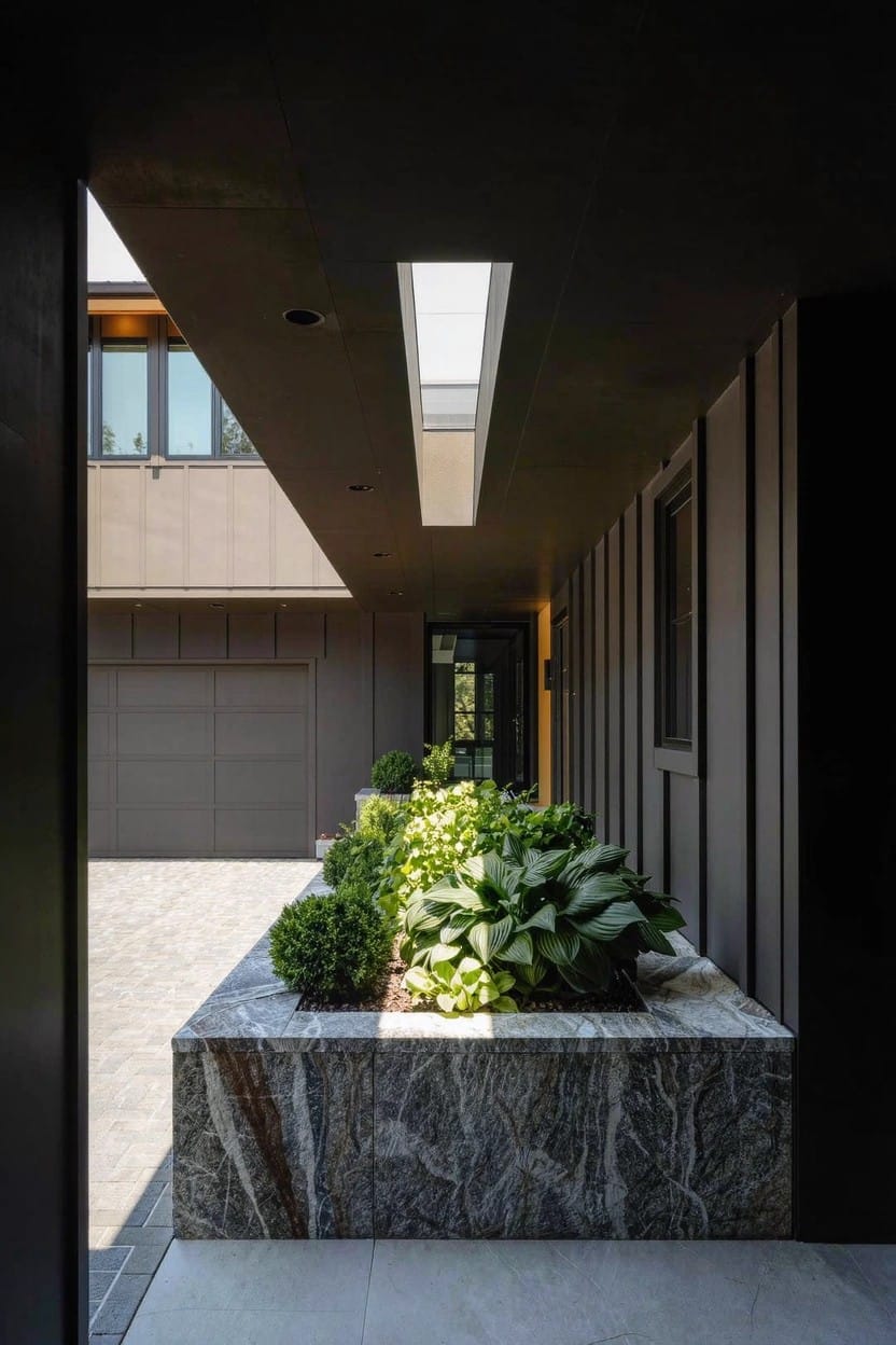 Modern home exterior with a covered walkway featuring a linear raised marble planter box filled with lush green shade plants like hostas along a dark vertical wood-clad wall, next to a gray garage door.