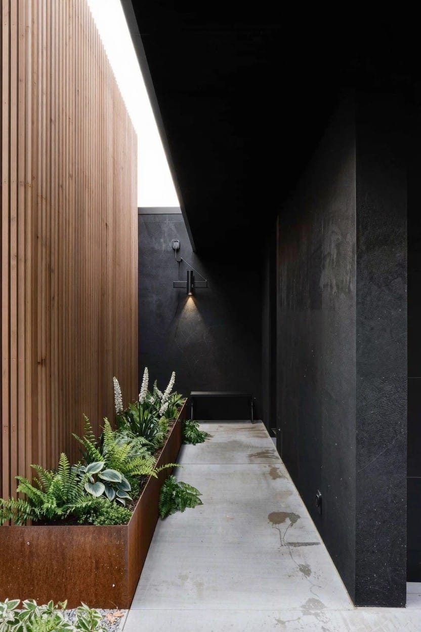 Narrow concrete pathway bordered by corten steel planters with ferns and green plants on one side and vertical wooden slats on the other, with dark walls, a bench, and wall lights.
