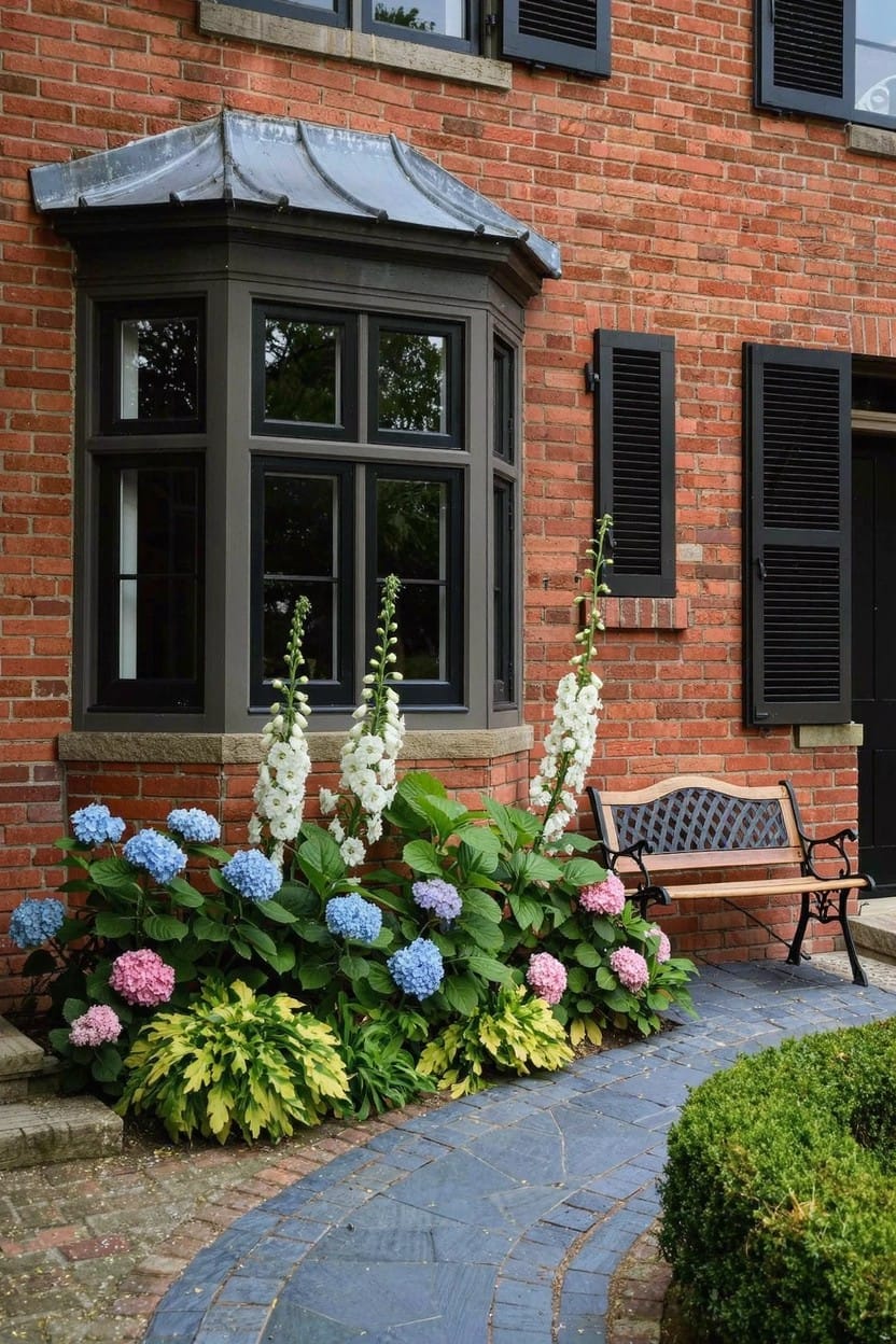 Brick house wall with bay window and black shutters, bordered by blue and pink hydrangeas, white foxgloves, green hostas, a wooden bench, and a curved stone path.