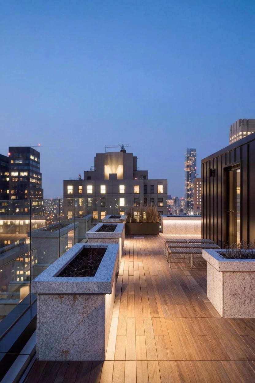 Rooftop terrace at dusk featuring large rectangular stone planters along glass railings, wooden deck flooring, metal seating, and surrounding city buildings.