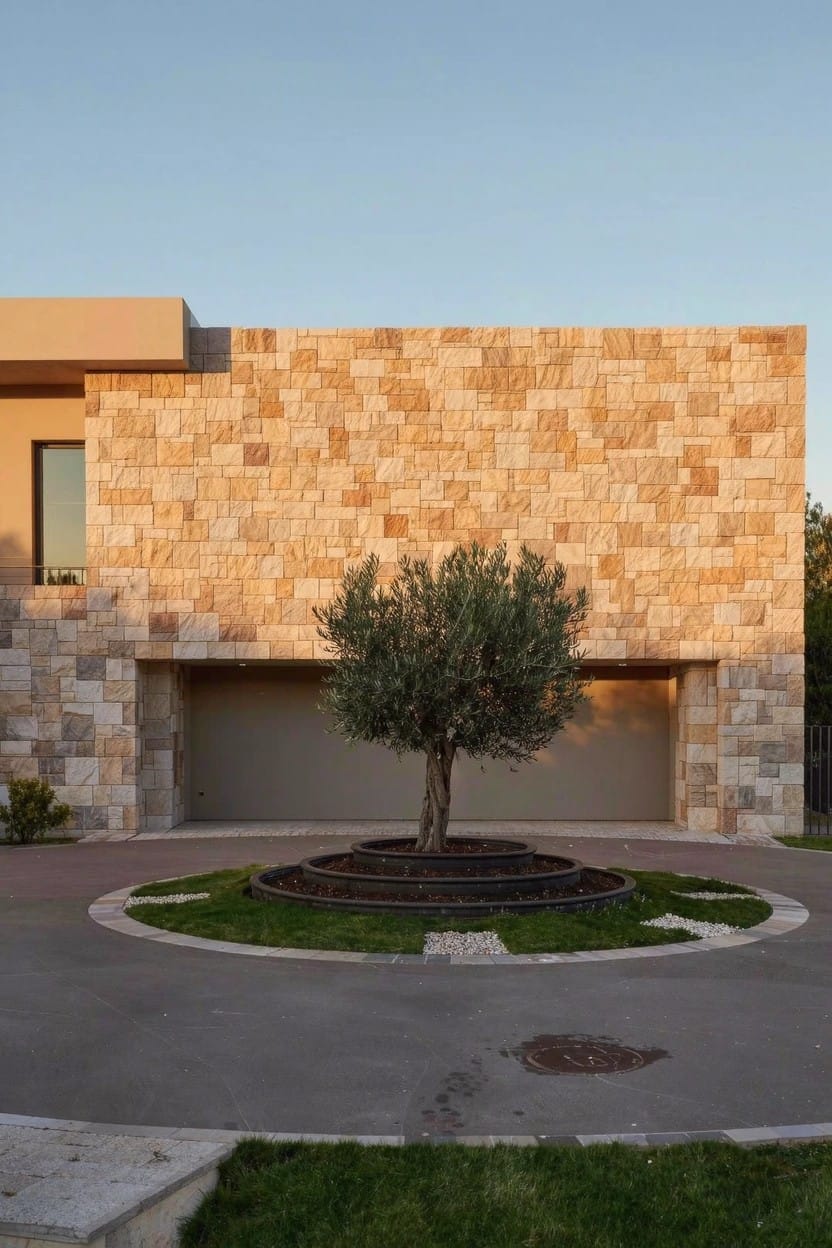 Beige stone house exterior with garage door and olive tree in circular raised gravel bed at driveway center.