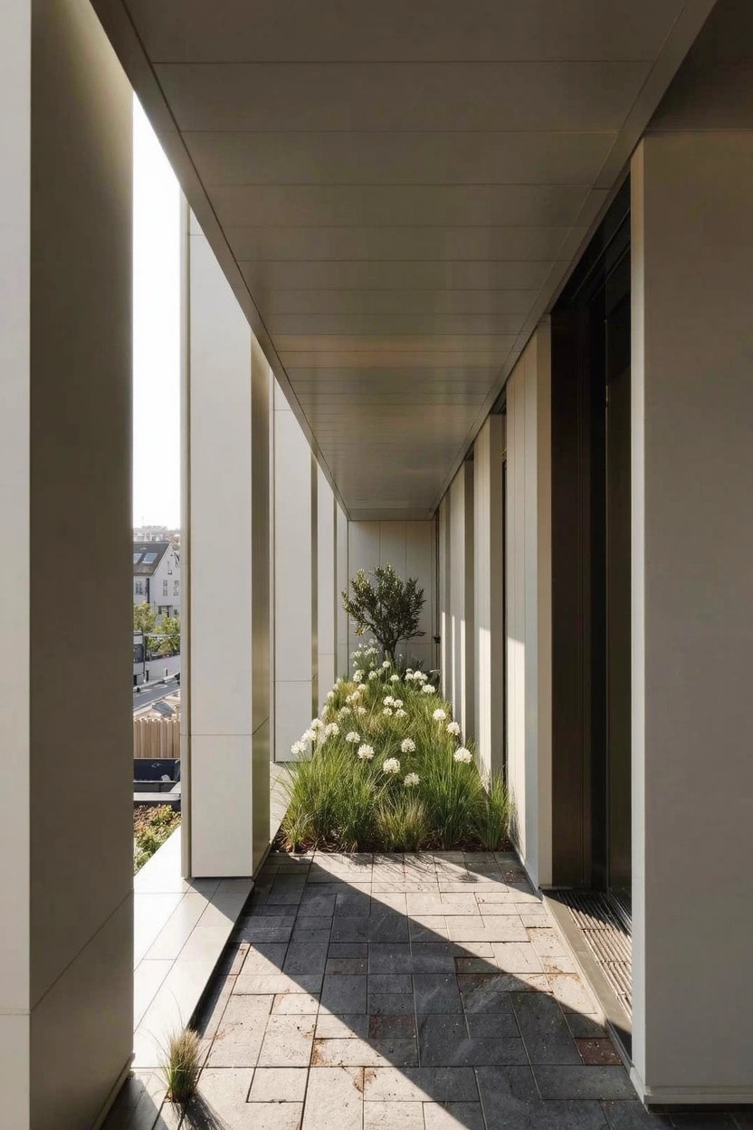 A long covered outdoor walkway flanked by tall white columns with a narrow planting bed of white flowers, grass, and a small tree along one side on stone pavers.