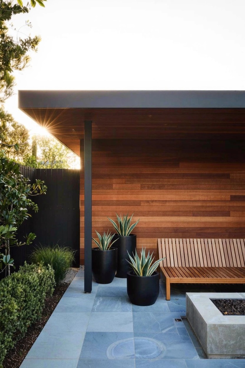 Outdoor patio with wooden bench, square fire pit, two large black pots holding agave plants, wooden siding wall, stone pavers, and nearby shrubs.