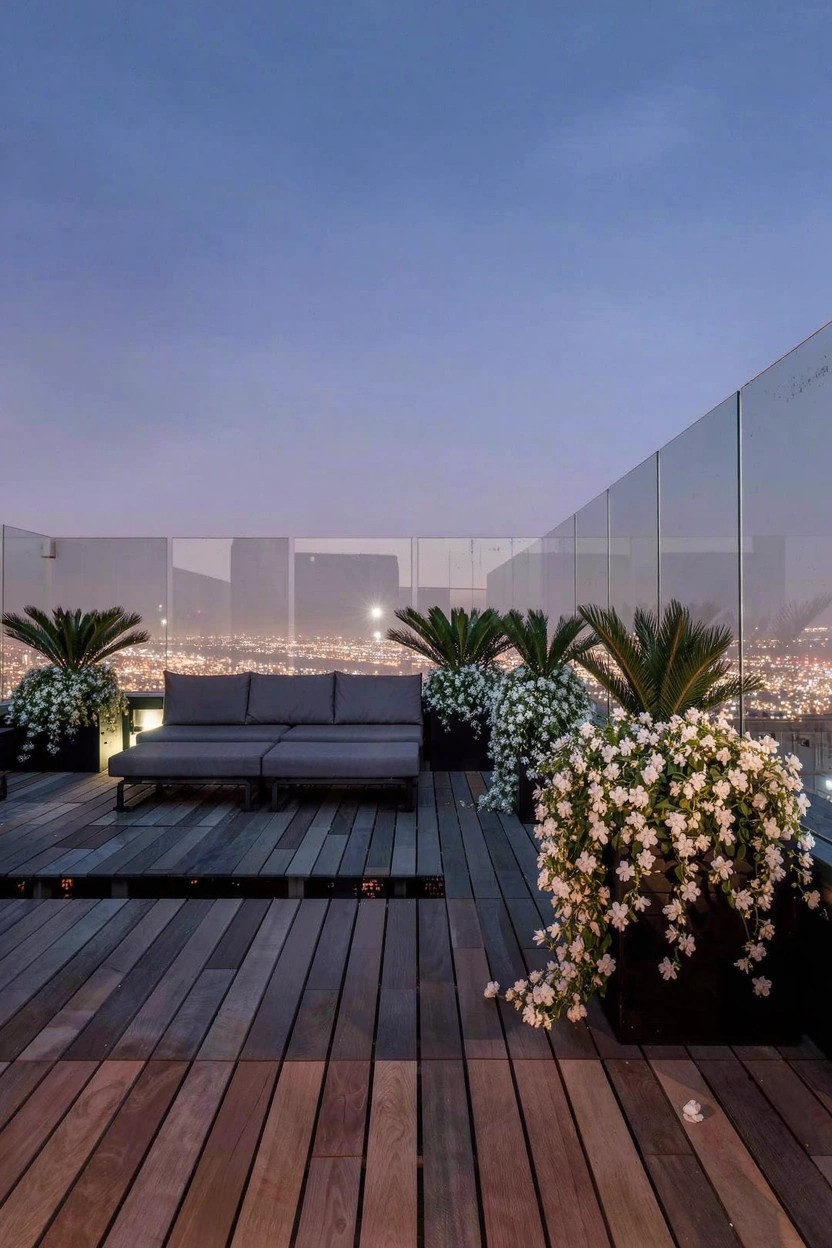 Rooftop patio at dusk with wooden deck, low glass railing, potted palms and large containers of white trailing flowers around a gray sofa and lamp, city lights visible beyond.