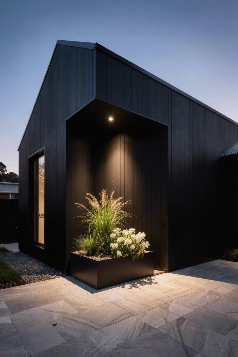 Black modern house exterior at dusk shows a recessed corner with vertical siding, a large black rectangular planter filled with tall grasses and white flowers, spotlight above, large window, and light gray stone pavers.