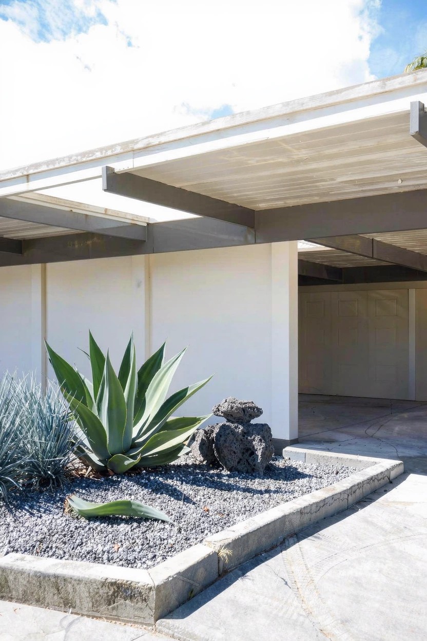 White modernist house with open carport and a raised corner bed containing large agave succulents, smaller blue agaves, rocks, and gravel mulch next to a concrete driveway.