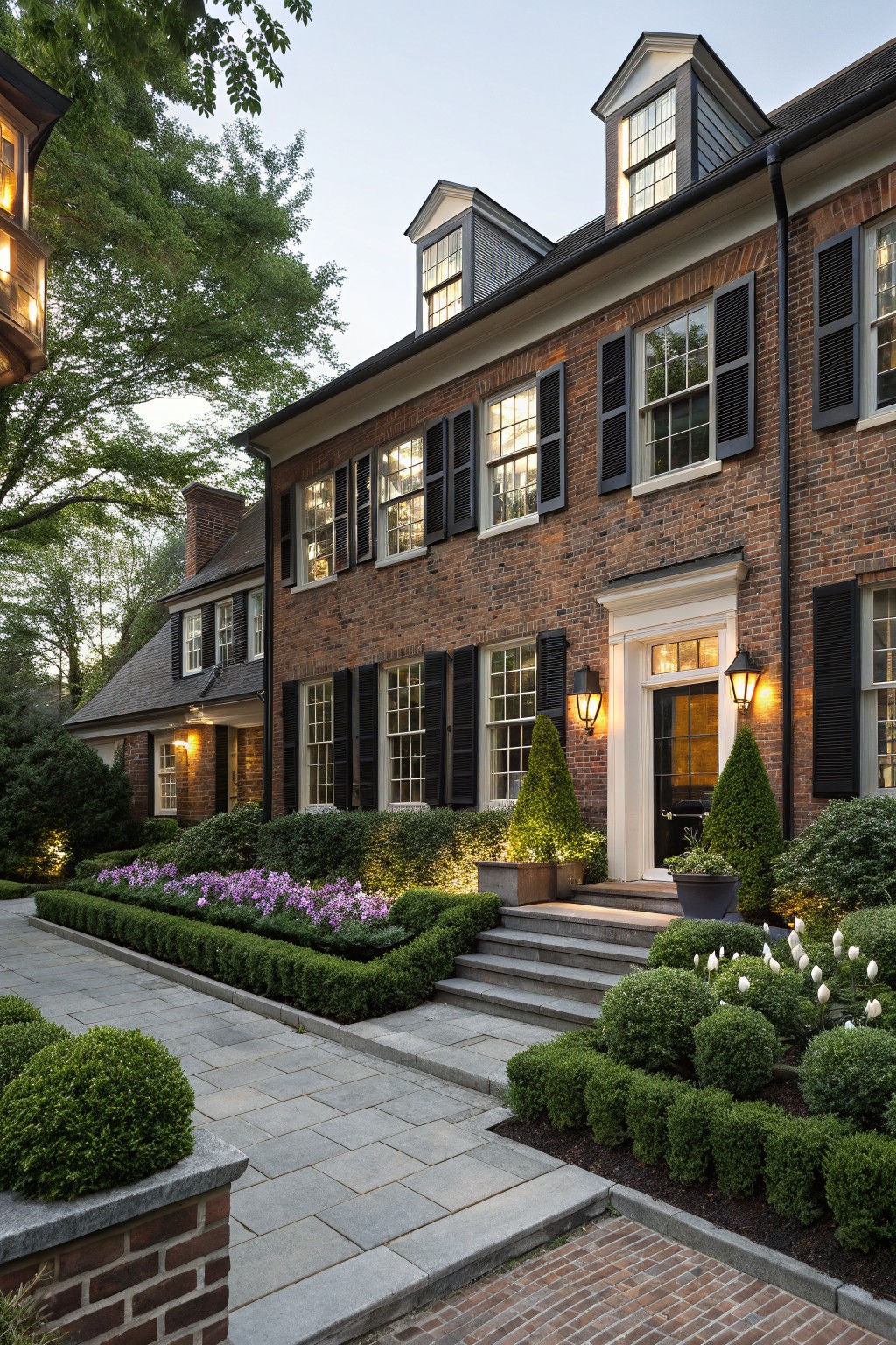 Red brick house exterior with black shutters and white trim, featuring manicured boxwood hedges bordering flower beds of purple flowers and white calla lilies along the foundation and slate pathway to the front door.