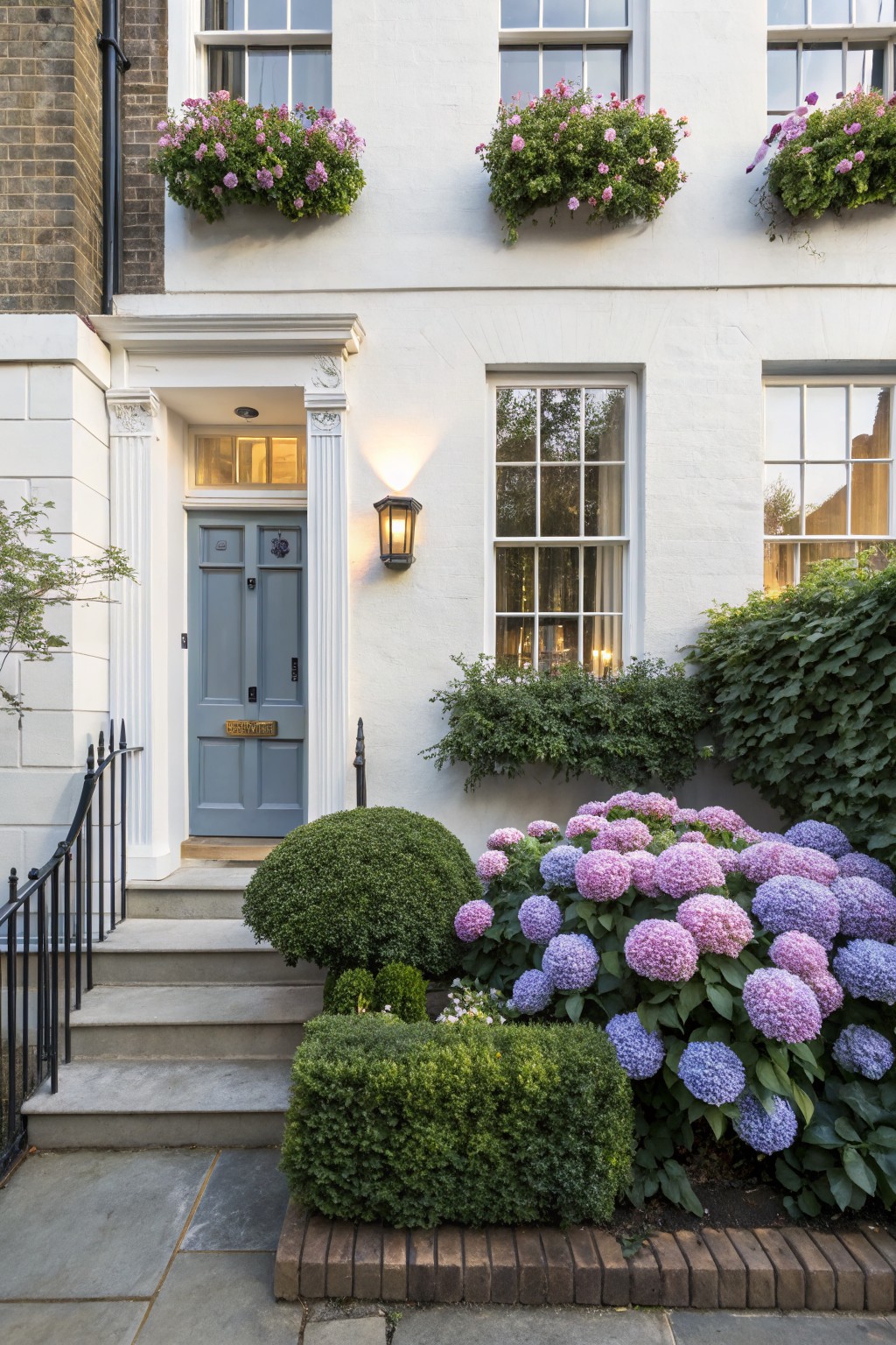 White terraced house exterior with blue front door, pink flower boxes on windows, purple and blue hydrangea beds, green boxwood shrubs, and brick edging along the front path.