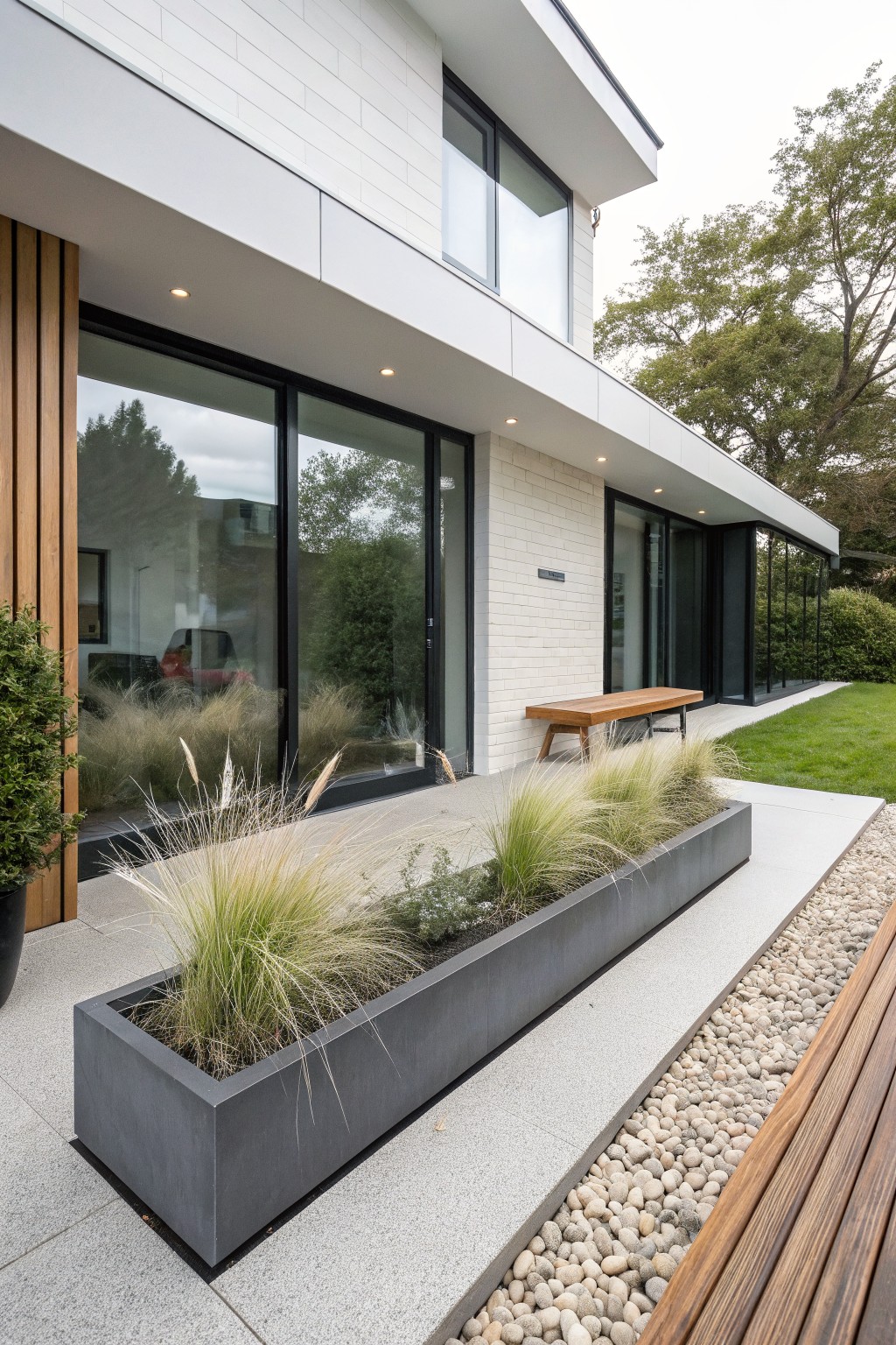 Side view of a modern white brick house with black-framed glass doors and windows, wooden bench on concrete paving, long black rectangular metal planter filled with tall ornamental grasses and low plants beside a pebble path and wooden decking.