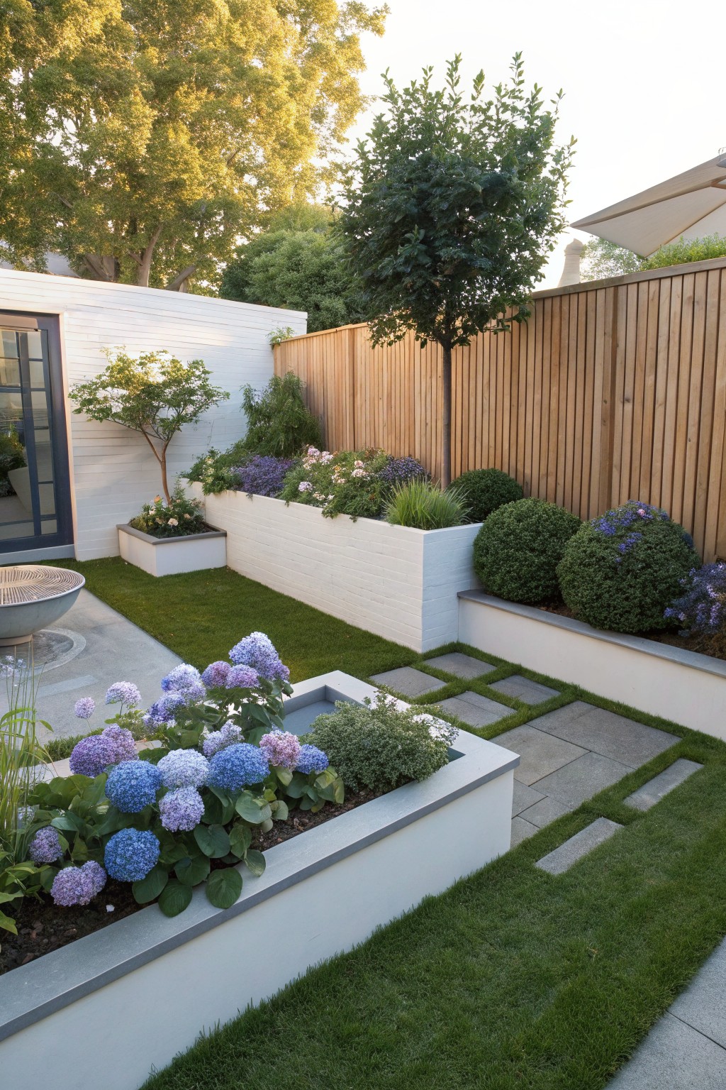 A backyard garden featuring raised white brick planters with blue hydrangeas and other flowers, surrounded by green grass, stone stepping paths, boxwood shrubs, a wooden fence, and trees.