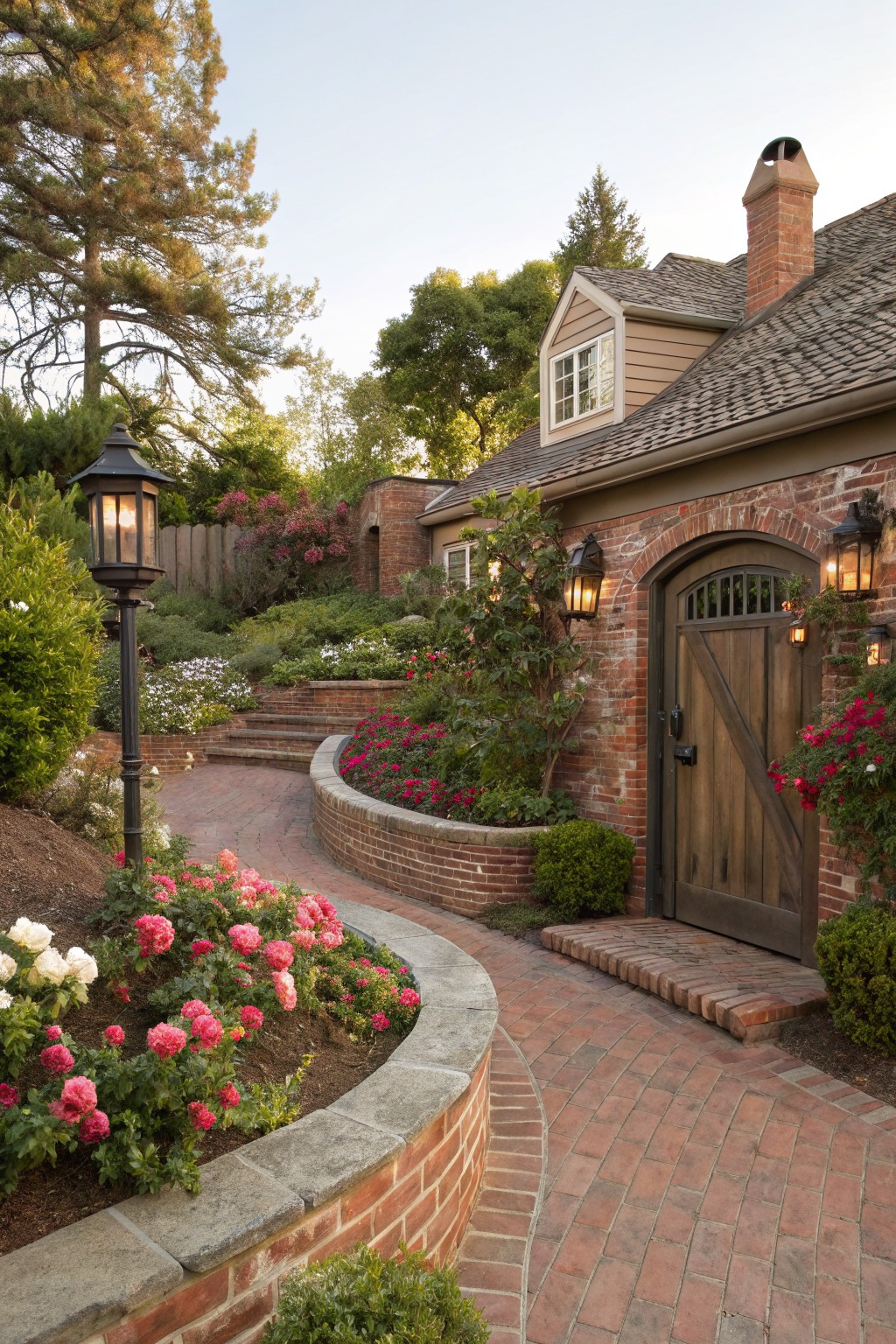 Curved brick retaining wall planted with pink and white roses beside a matching brick pathway leading uphill to an arched wooden door on a brick house exterior, flanked by lanterns and garden shrubs.
