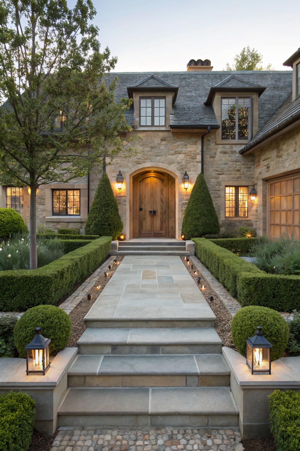 Stone house facade with arched wooden entry door, wide flagstone pathway lined with low-voltage lights, bordered by clipped boxwood hedges and topiary trees, flanked by lanterns and steps.