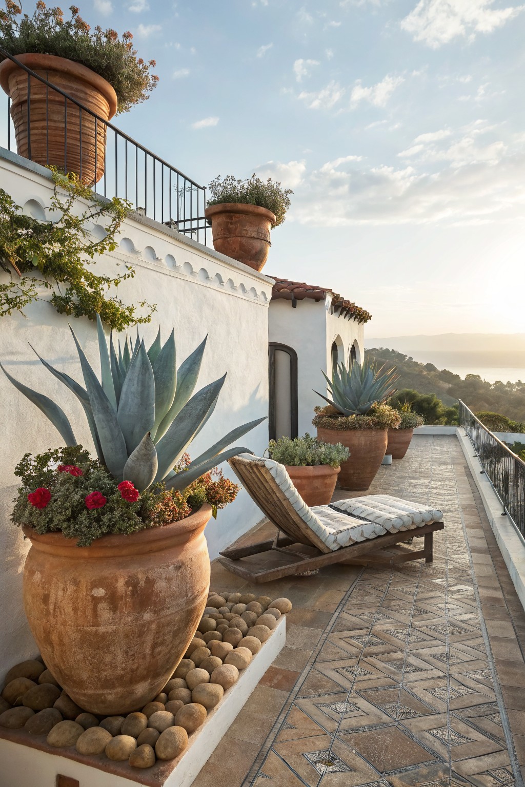 White stucco balcony terrace with large terracotta pots of blue agave succulents and red roses, a wooden chaise lounge, pebble border, and distant ocean view at sunset.