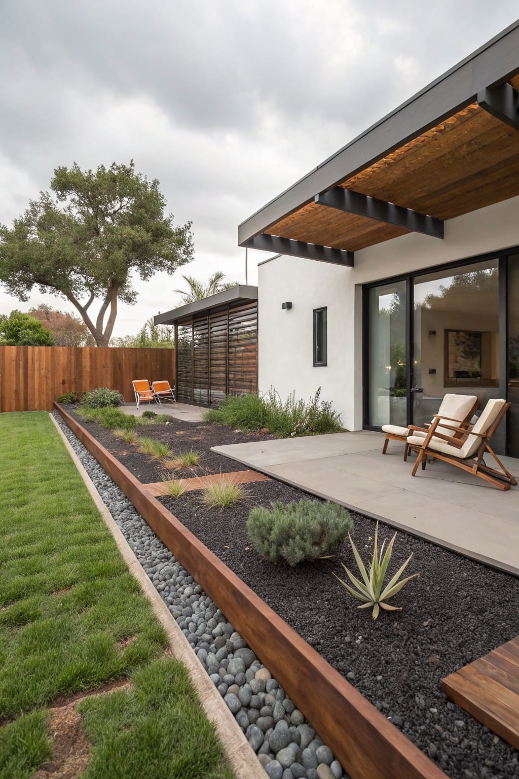 Modern white house exterior with concrete patio, wooden lounge chairs, and adjacent garden beds edged by horizontal wood boards containing black gravel mulch, succulents, and agave plants, bordered by green lawn and pebble strip.
